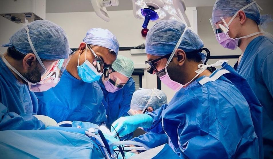 A team of plastic and reconstructive surgeons operating a patient who cannot be seen. The surgeons are wearing blue scrubs, surgical hats and masks.