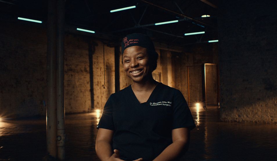 Portrait of Maryam Alfa-Wali, trauma surgeon at St Mary's Hospital dressed in hospital scrubs and smiling towards the camera.