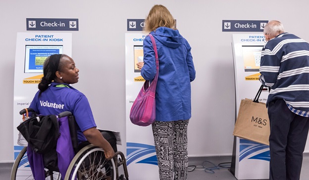 Two patients checking in at a hospital reception. A volunteer in a wheelchair is assisting both patients.