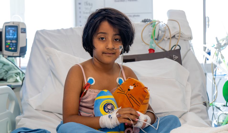 Photo of a young girl sitting in a hospital bed. She is cuddling some teddies and looking towards the camera. Various wires can be seen behind her and connected to her body.