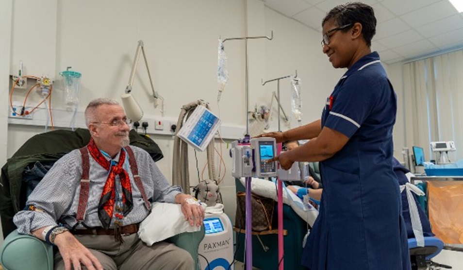 A male patient sits in an armchair on a chemotherapy ward at Imperial College Healthcare while a nurse adjust his chemotherapy treatment.