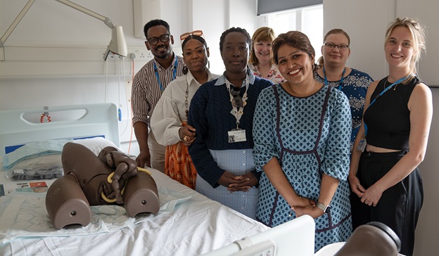 Photograph of Imperial College Healthcare NHS staff standing next to a hospital bed which has a darker skin tone manikin on it.