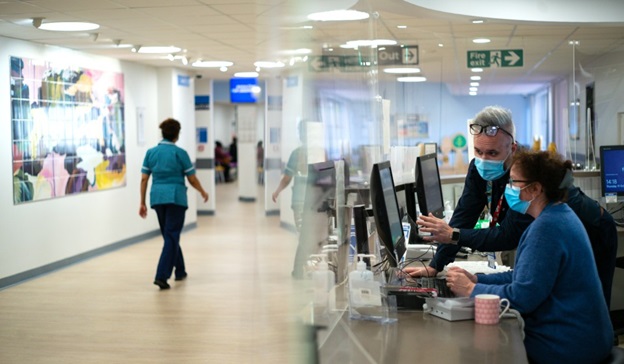 A health care worker walks through a hospital corridors while two others work at a computer