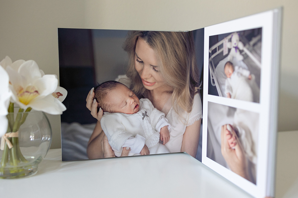 Open photo album on table next to a vase of white flowers. The photos in the album are of a newborn. The main photo shows the mother holding baby in her arms. 