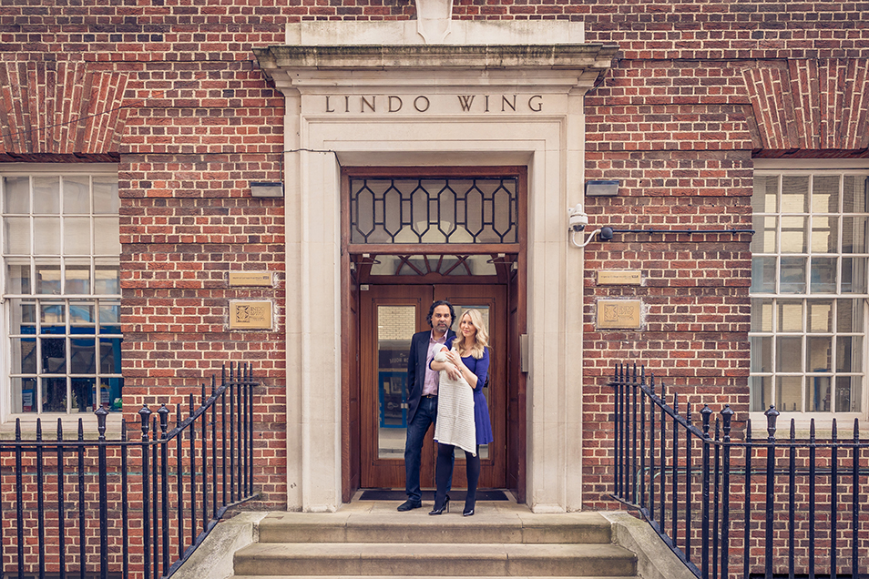 Mother, father and newborn being held in mother’s arms while smiling and staring into the camera as they standi outside the entrance to the Lindo Wing hospital.