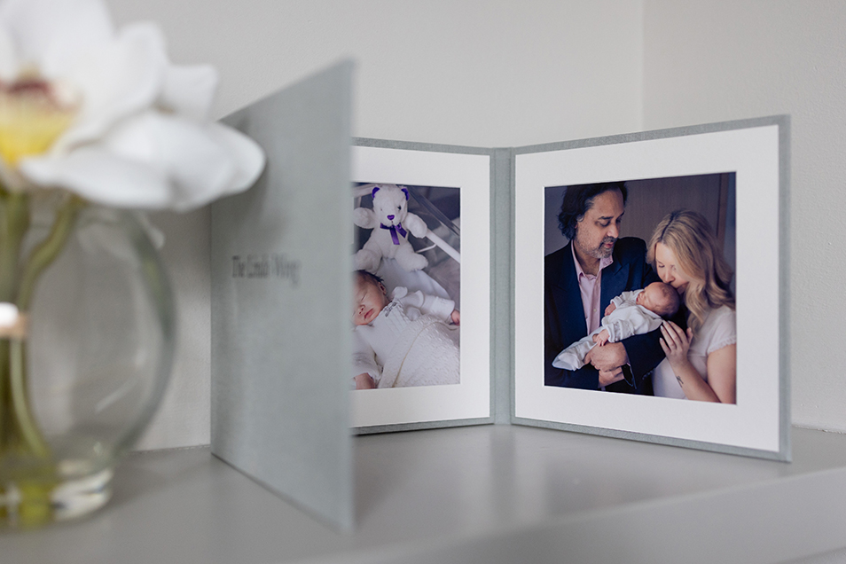 Open photo album on a table showing a photo of a mother, father and baby. The baby is being cradled in father’s arms as mother kisses baby on forehead.